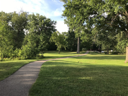 A winding path through a lush green park with trees and a glimpse of a bridge in the background.