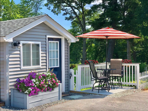 A cozy gray cottage with a white porch, red-striped umbrella, and colorful flower boxes in a sunny outdoor setting.
