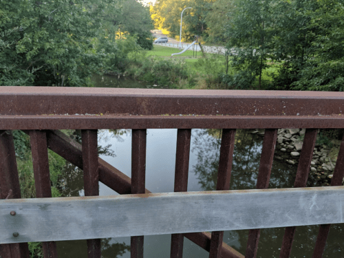 A rusty metal bridge railing overlooking a calm river surrounded by trees and a road in the background.