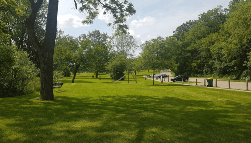 A grassy park with trees, a swing set, and a parking area in the background under a partly cloudy sky.