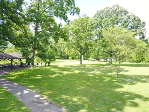 A sunny park scene with green grass, trees, and a gazebo, featuring people walking in the distance.