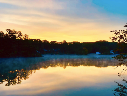 A serene lake at sunrise, with mist rising and colorful reflections of trees and sky on the water's surface.