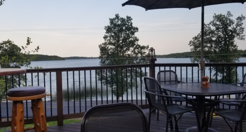 A serene lakeside view from a wooden deck with tables and chairs, surrounded by trees under a clear sky.