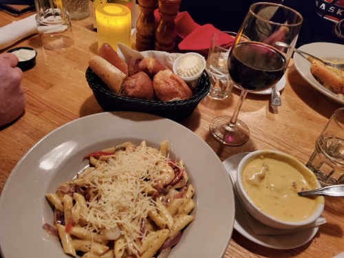 A plate of pasta topped with cheese, a bowl of soup, a glass of red wine, and a basket of bread rolls on a wooden table.