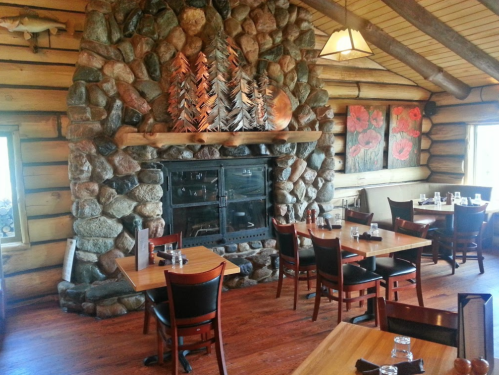 Cozy restaurant interior with wooden decor, stone fireplace, and tables set for dining. Warm, rustic atmosphere.