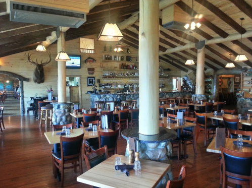 Cozy restaurant interior with wooden beams, stone accents, and tables set for dining. Bar area in the background.