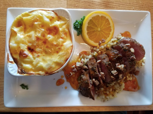 A plate featuring sliced meat with sauce, a side of creamy potato dish, and a slice of orange on a bed of grains.