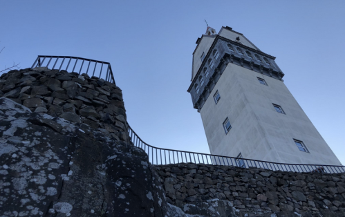 A tall tower stands on a rocky hill, surrounded by a metal railing against a clear blue sky.