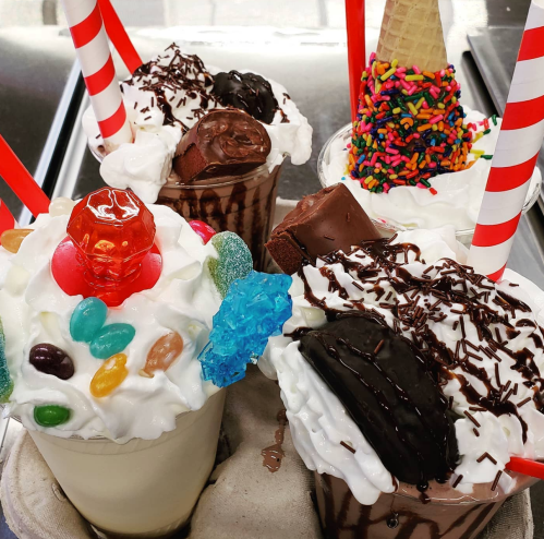 Four colorful milkshakes topped with whipped cream, candy, and sprinkles, served with red straws in a fun presentation.