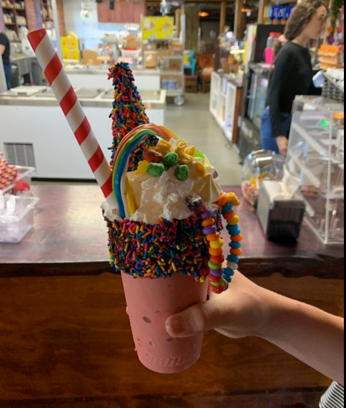 A colorful milkshake topped with whipped cream, sprinkles, a rainbow candy, and a striped straw, held in a hand.