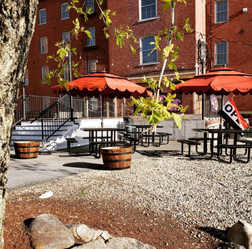 Outdoor seating area with red umbrellas, picnic tables, and a gravel path near a brick building.