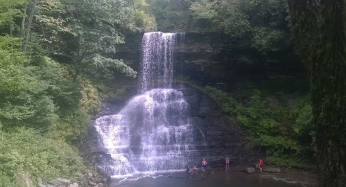 A serene waterfall cascades down rocky cliffs, surrounded by lush greenery and people enjoying the water below.