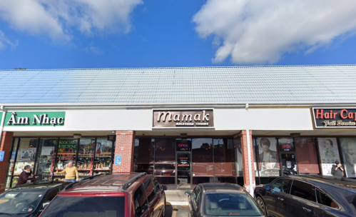 A storefront with the sign "Mamak" in a shopping plaza, flanked by other businesses and parked cars in front.