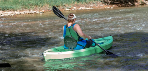 A person paddles a green kayak on a calm river, wearing a blue life jacket and a headband.