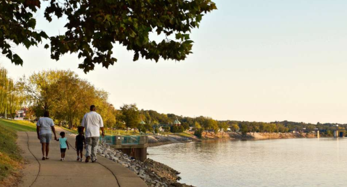 A family walks along a riverside path, enjoying a peaceful evening by the water. Trees line the shore in the background.
