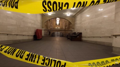 Caution tape blocks off a dimly lit hallway in a public building, with furniture covered in cloth in the background.