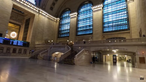 Interior of a grand train station with high ceilings, large windows, and a staircase leading to ticket counters and tracks.