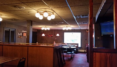 Interior of a dimly lit restaurant with wooden paneling, round lights, and empty tables. A TV is visible in the corner.