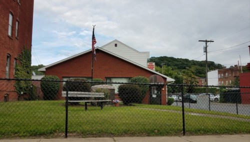 A red brick building with a flagpole, surrounded by a green lawn and a chain-link fence, under a cloudy sky.