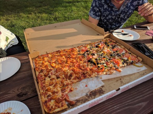 A large pizza box with half pepperoni and half veggie pizza, placed on a wooden table outdoors.