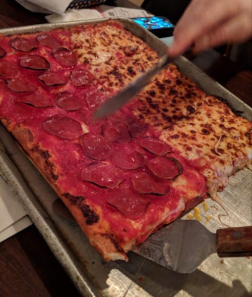 A tray of pizza with one half topped with pepperoni and the other half with cheese, being sliced with a knife.