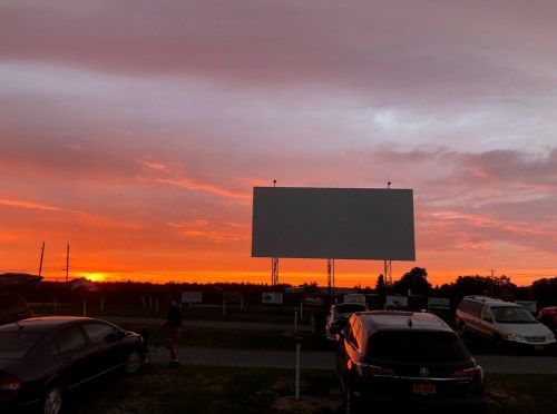 A vibrant sunset with orange and pink hues behind a blank outdoor movie screen and parked cars.