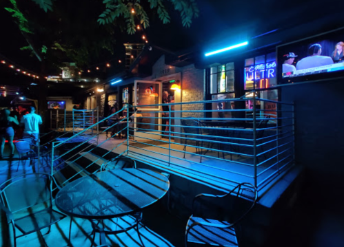 A dimly lit outdoor bar area with blue neon lights, tables, and people socializing in the background.