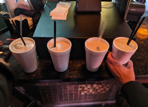 Four white cups with straws are lined up on a counter, with hands holding one cup.