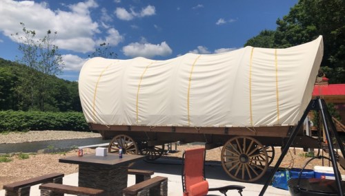 A covered wagon on wheels sits outdoors near a river, with a blue sky and clouds in the background.