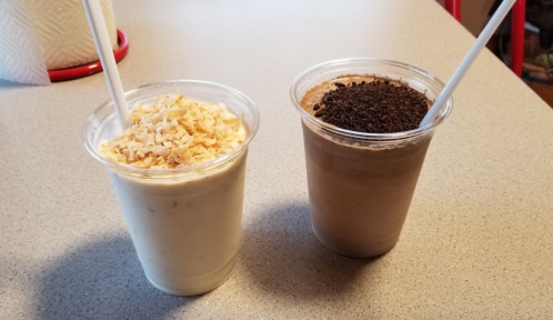 Two cups of smoothies on a countertop: one topped with shredded coconut and the other with crushed cookies.