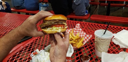 A hand holding a large burger with lettuce and tomato, alongside a side of fries and a drink on a red table.