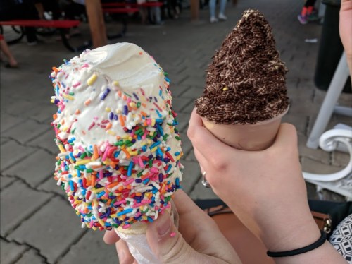 Two hands hold ice cream cones: one with colorful sprinkles and the other topped with chocolate sprinkles.
