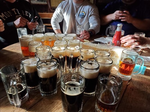 A table filled with various beer mugs and glasses, with people’s hands and phones visible in the background.