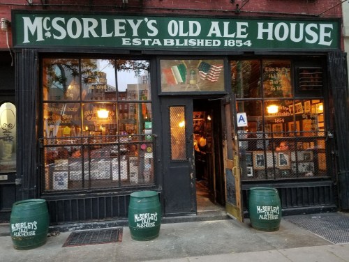 Exterior of McSorley's Old Ale House, a historic bar established in 1854, featuring green barrels and warm lighting.