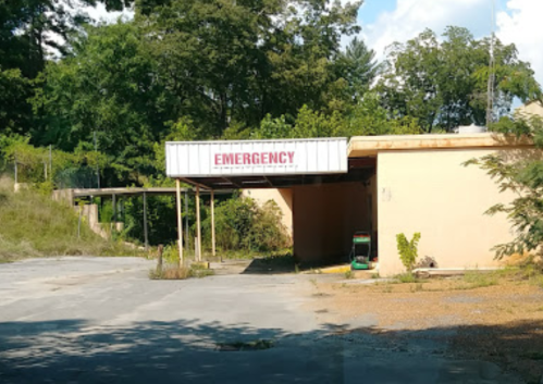 Abandoned building with a sign reading "EMERGENCY" and overgrown vegetation in the surrounding area.