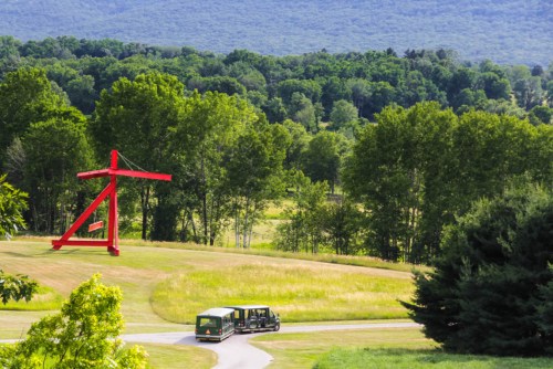 A bright red sculpture stands in a green landscape, with a tour vehicle winding through the grassy area.