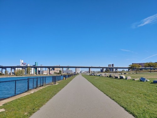 A clear blue sky over a pathway beside a river, with a bridge and buildings in the background. Green grass and rocks line the path.