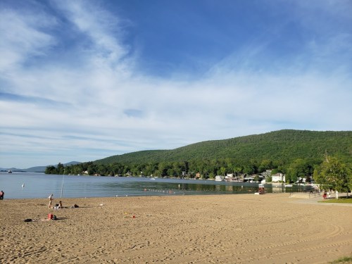 A serene beach scene with a sandy shore, calm water, and green hills under a blue sky with scattered clouds.
