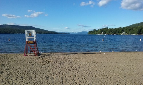 A sandy beach with a lifeguard stand, calm water, and distant mountains under a clear blue sky.