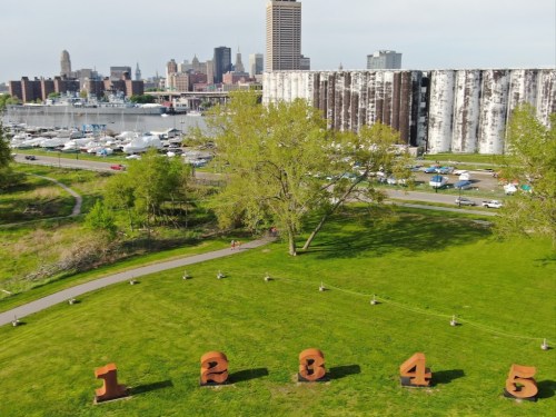 Aerial view of large numbered sculptures on green grass, with a city skyline and marina in the background.