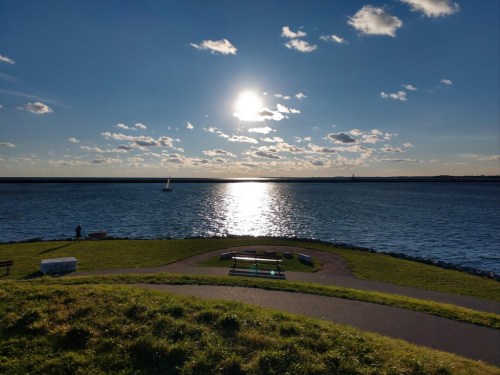 A serene waterfront scene with a shining sun, clouds, and a sailboat on the water, surrounded by green grass and benches.