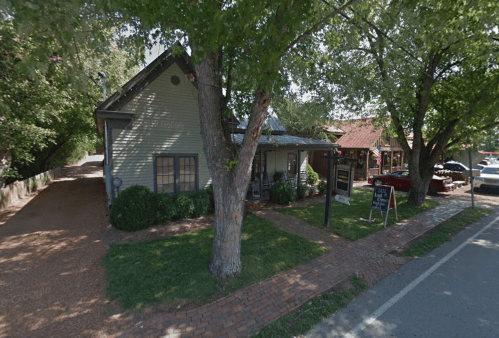 A quaint, green house with a porch, surrounded by trees and a gravel path, near a street with parked cars.