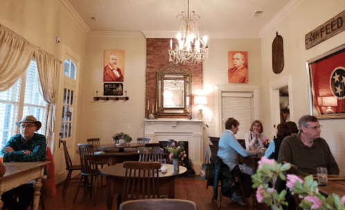 Cozy café interior with wooden tables, patrons chatting, and vintage portraits on the walls. Flowers on tables.