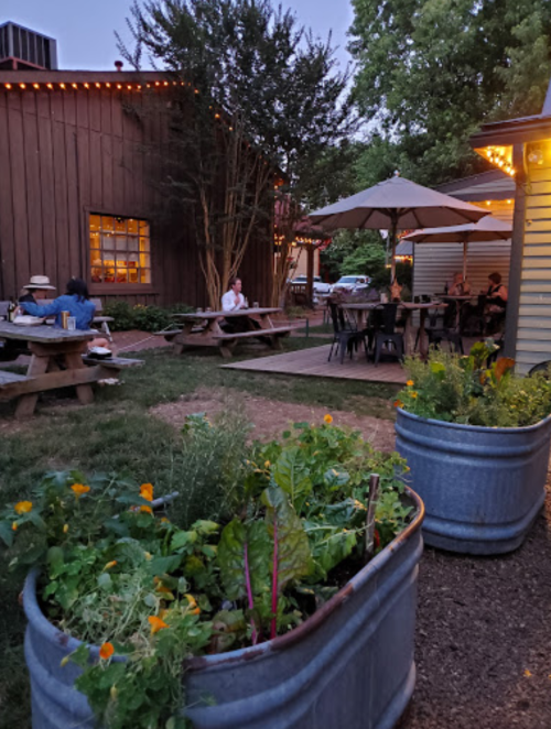A cozy outdoor dining area with picnic tables, umbrellas, and planters filled with greenery, illuminated by string lights.