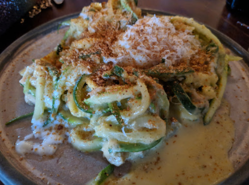 A plate of creamy zucchini noodles topped with grated cheese and breadcrumbs, garnished with herbs.