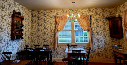 Cozy dining room with floral wallpaper, wooden tables, chairs, and a chandelier, featuring a window with drapes.