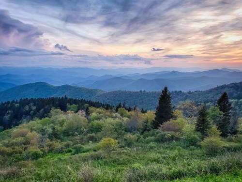 A scenic view of rolling mountains under a colorful sunset, with lush greenery in the foreground.