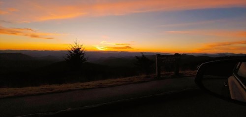 Sunset view from a mountain overlook, with rolling hills and a colorful sky in the background. Car door visible on the right.