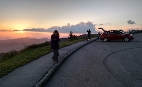 A woman walks along a scenic overlook at sunset, with a car parked nearby and another person in the background.