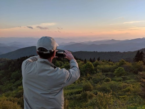 A person in a cap takes a photo of a scenic mountain view at sunset, surrounded by lush greenery.